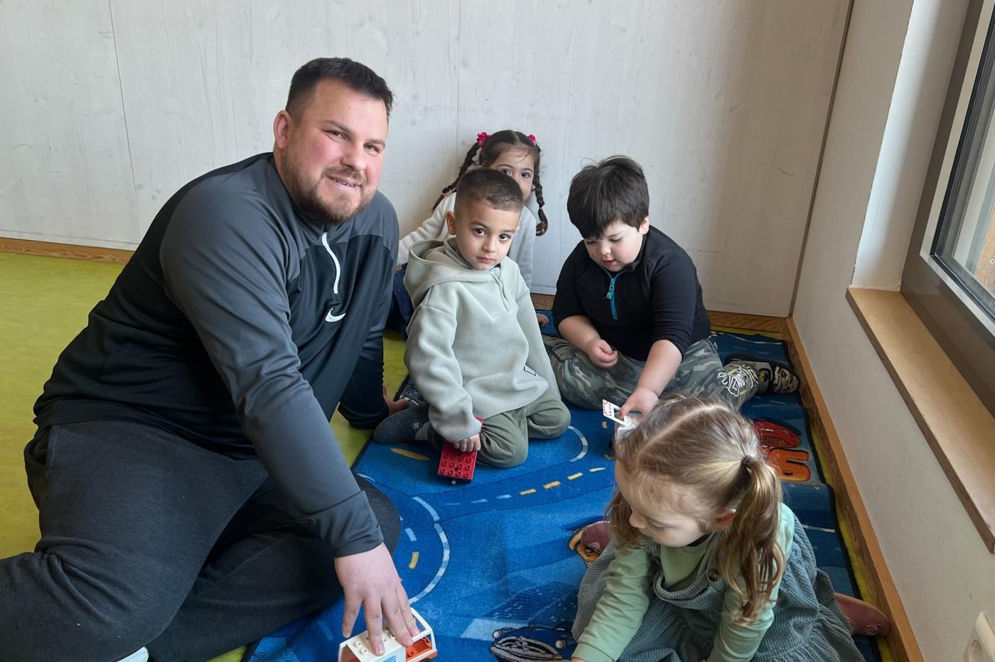 Stadtverwaltung Ein Mann spielt mit Kindern auf einem Teppichboden mit Autos