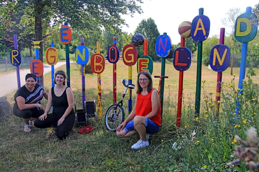 Drei Frauen hocken vor einigen bunten Stangen, an denen Buchstaben das Wort Regenbogenland ergeben