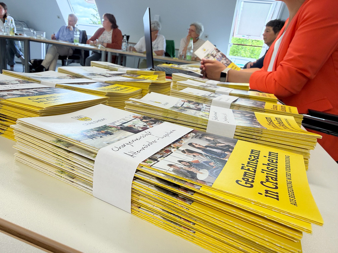 Einige Flyer-Pakete liegen auf einem Tisch, im Hintergrund sind Menschen zu erkennen