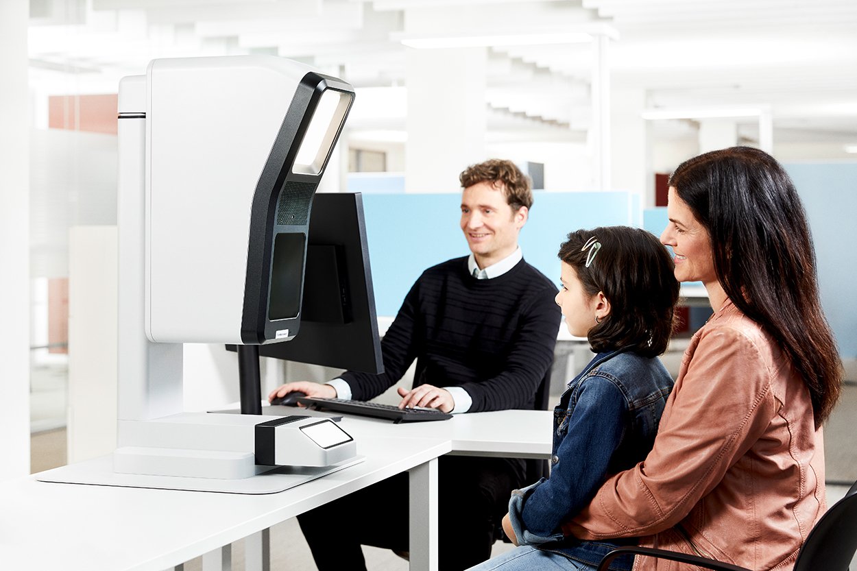 Eine Frau mit Kind auf dem Schoß sitzt vor einem Foto-Gerät, das ein Mann im Hintergrund bedient.