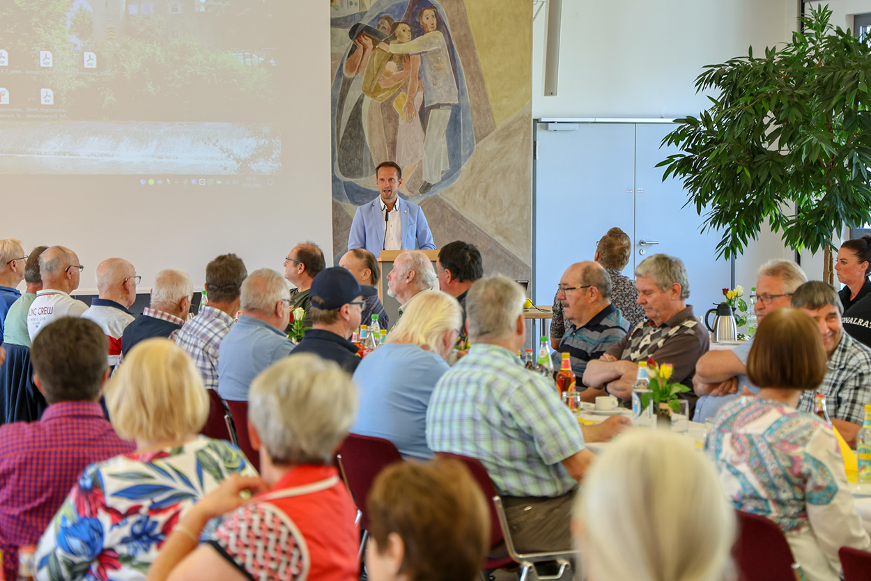 Ein Mann im Hintergrund am Rednerpult, davor viele Zuhörende an Tischen