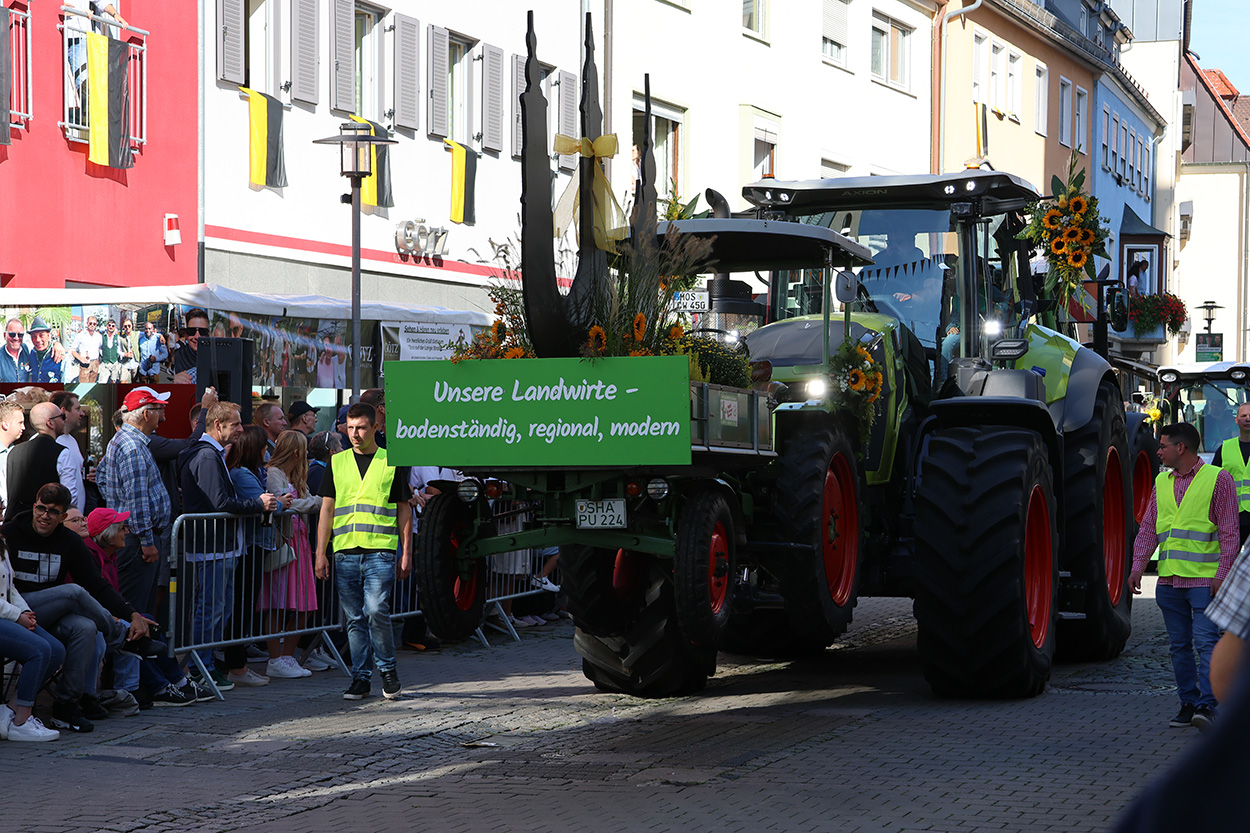 Ein großer Zraktor mit Schild vorne dran begleitet von Männern in gelben Westen, am Rand Zuschauendeviel