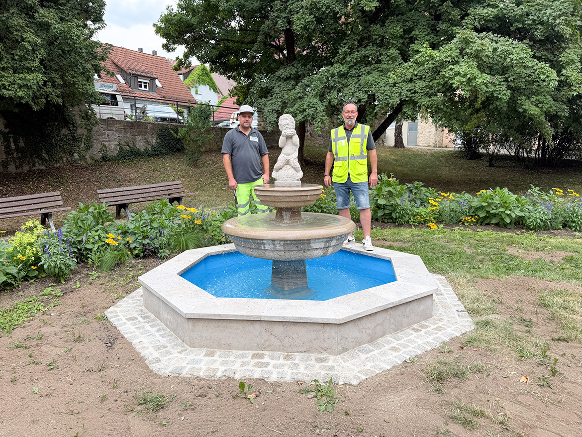 zwei Männer stehen an einem Brunnen mit Figur oben drauf