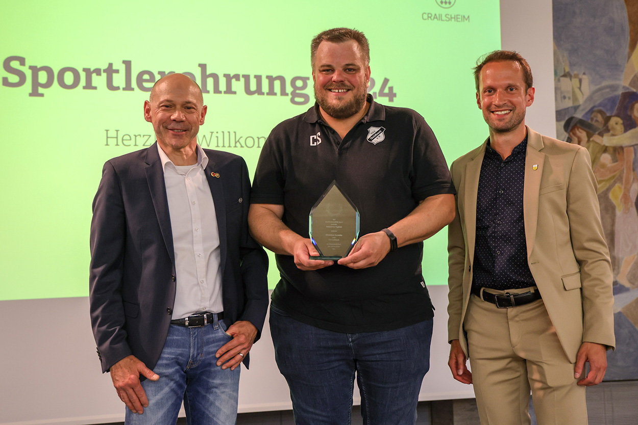 drei Männer lächelnd in die Kamera, der Mittlere hält eine Auszeichnung in der Hand