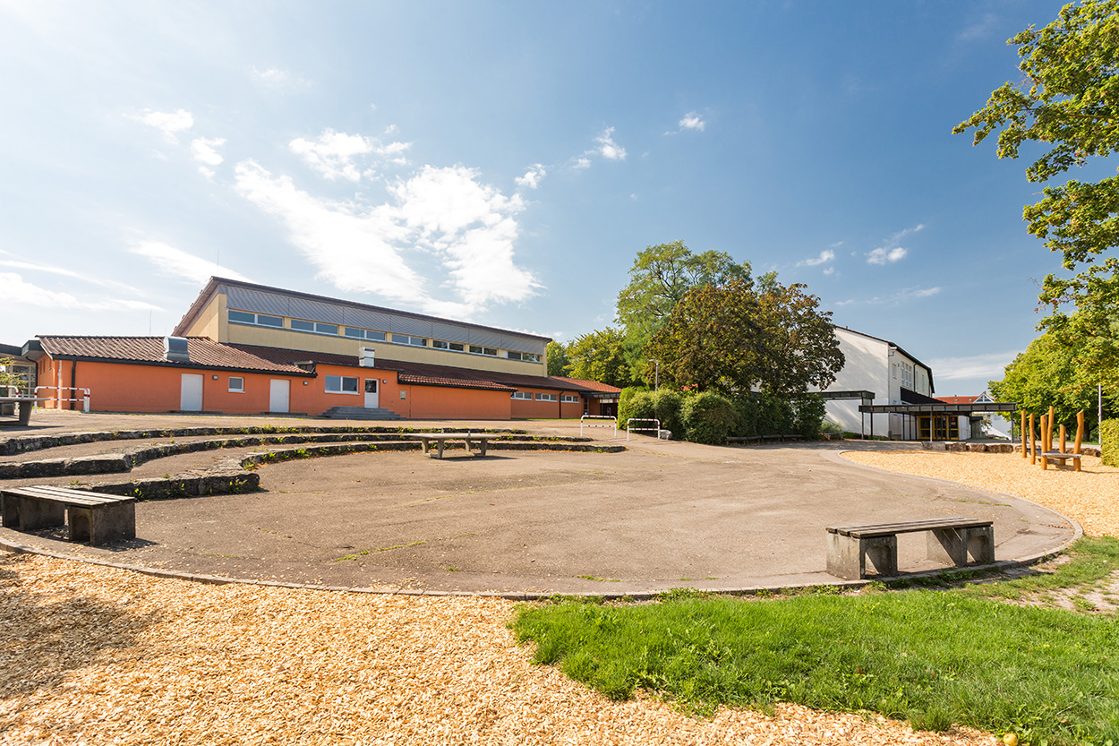ein großer Platz mit Bank und angrenzendem Spielplatz, dahinter ein Schulgebäude