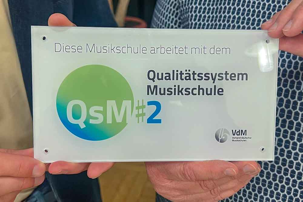 Eine kleine Tafel wird in die Kamera gehalten, auf der steht u.a. Qualitätssystem Musikschule