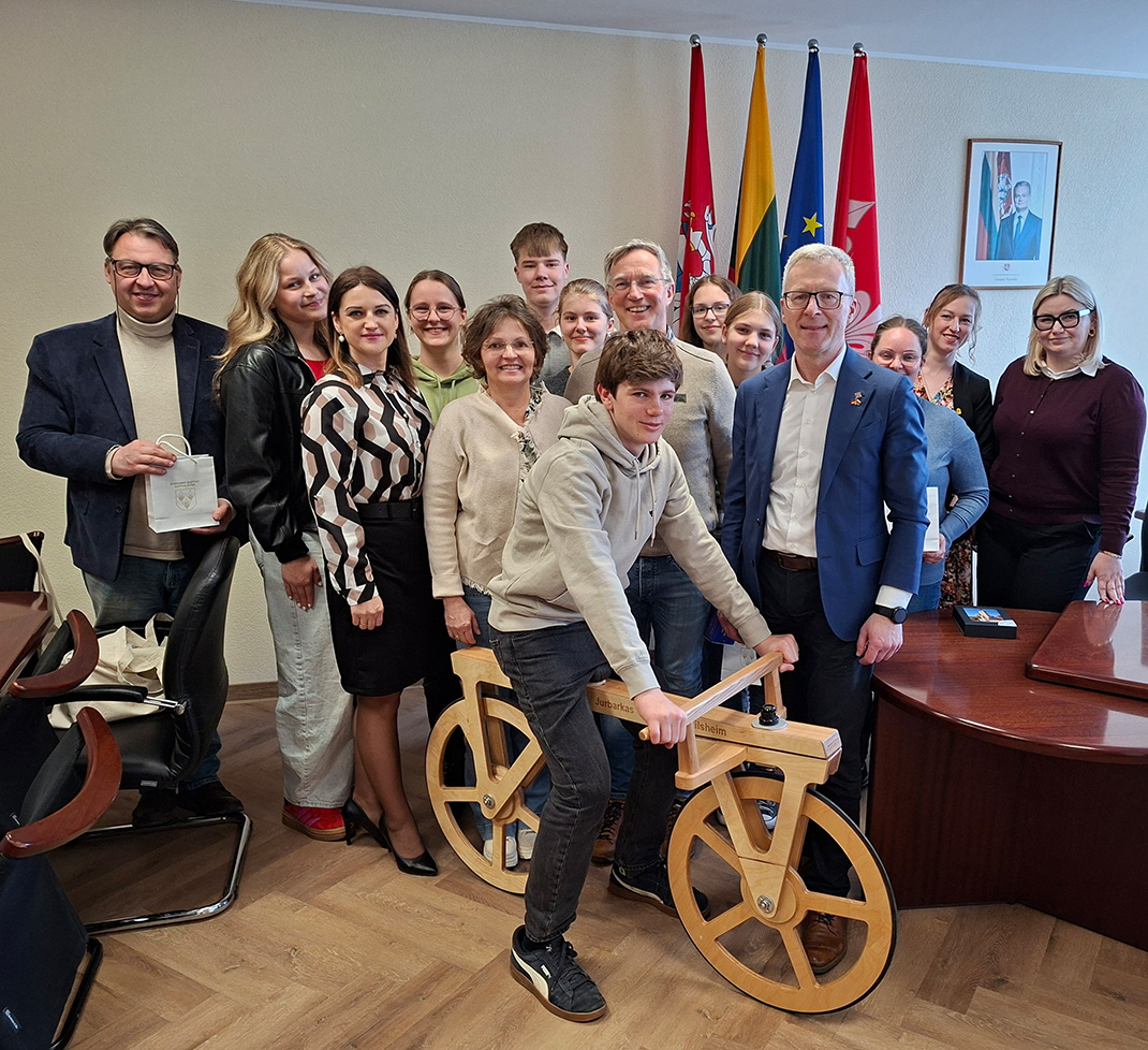 Mehrere Personen stehen in einem Büro, ein junger Mann sitzt auf einem Draisrad