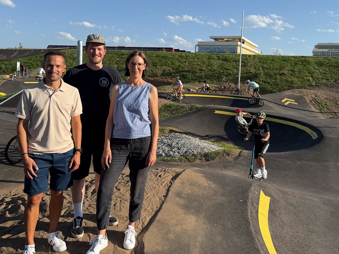 Zwei Männer und eine Frau stehen am Rand eines Pumptracks und lächeln in die Kamera