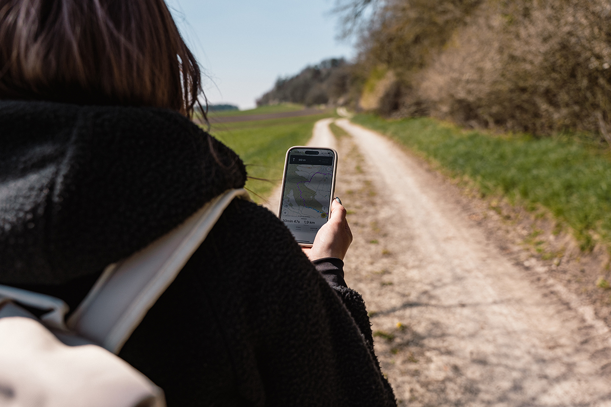 Eine Wanderin hat ein Handy in der Hand und hat einen Feldweg vor sich