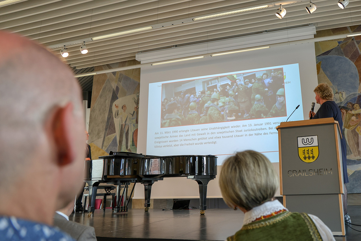 Eine Frau an einem Rednerpult auf eienr Bühne blickt auf die Leinwand hinter sich, im Vordergrund Köpfe von Zuschauern.