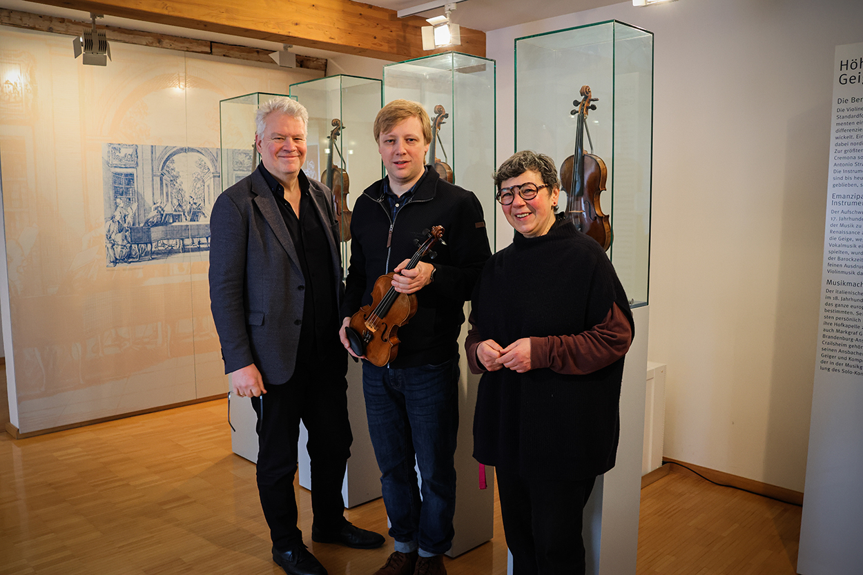Zwei Männer und eine Frau stehen mit einer Geige vor einer Vitrine