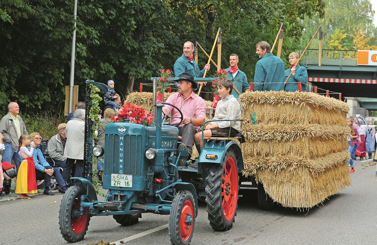 Ein Traktor zieht einen Hänger mit Strohballen, darauf einige Männer