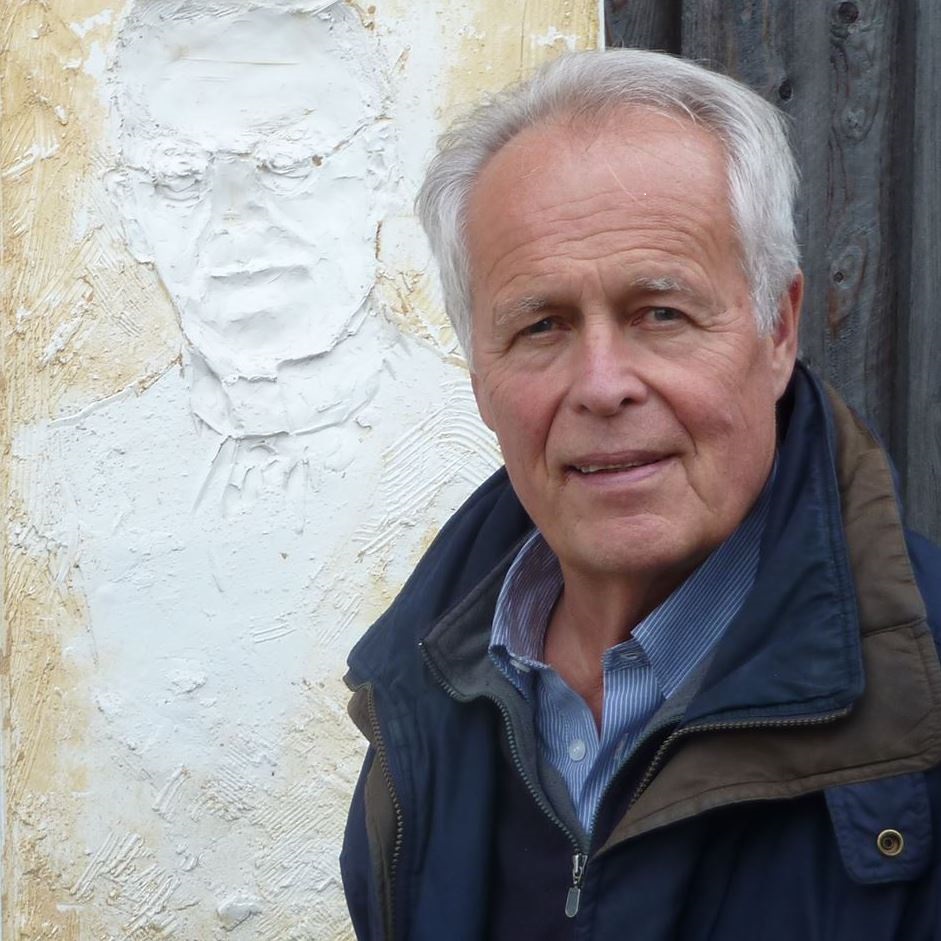 Ein älterer Herr blickt neben einem Bildnis an der Wand in die Kamera