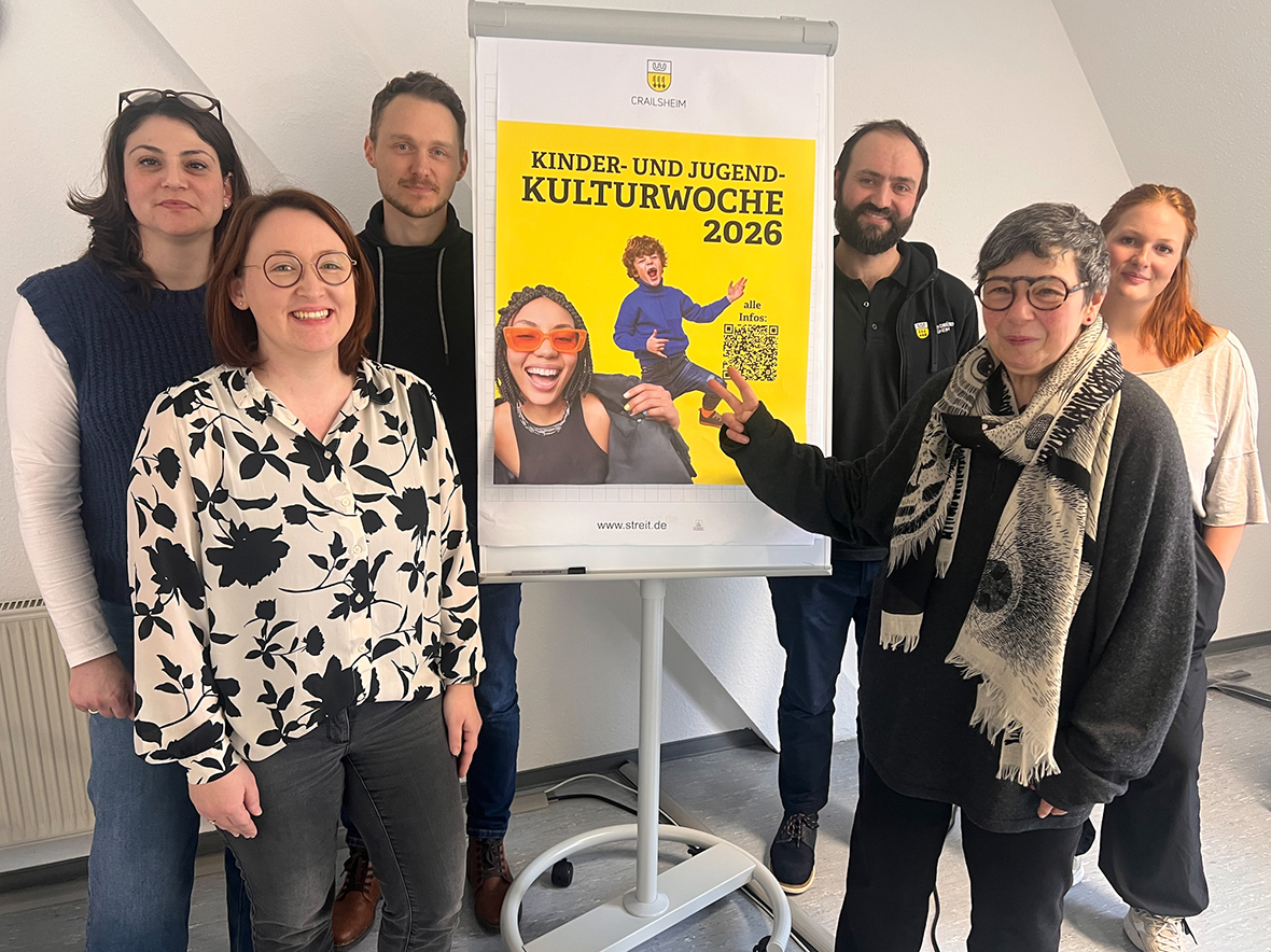 Einge sechsköpfige Gruppe steht lächelnd um ein buntes Plakat