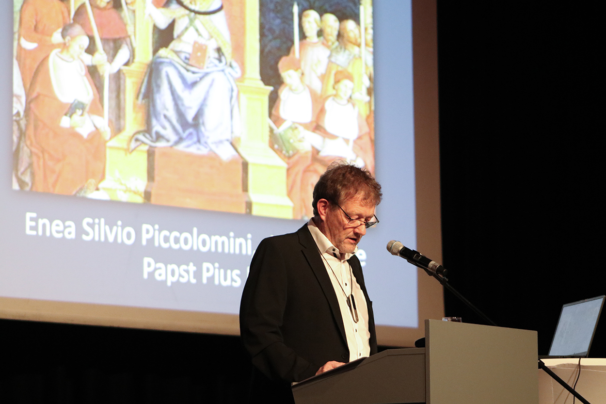 ein Mann mit Brille steht an einem Rednerpult vor einer Leinwand
