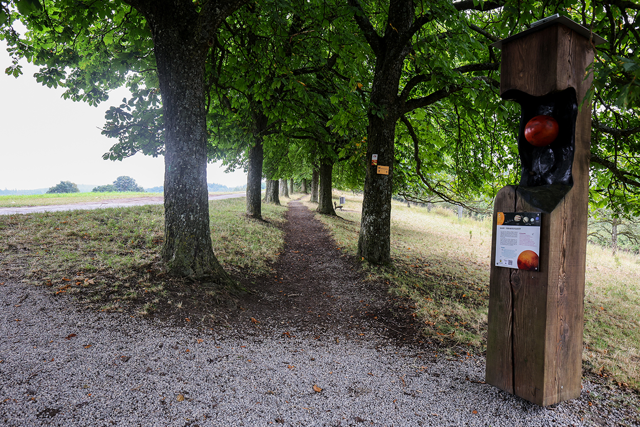 eine Baumreihe mit einem Kiesweg, daneben eine Stele mit einem Planeten