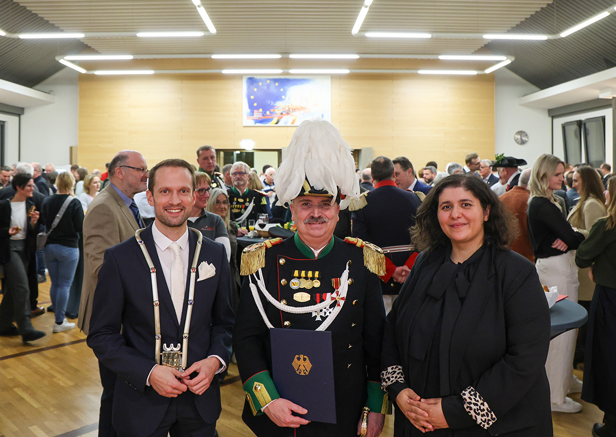 Ein Mann in Gardeuniform steht zwischen einem Mann und einer Frau, alle lächeln in die Kamera
