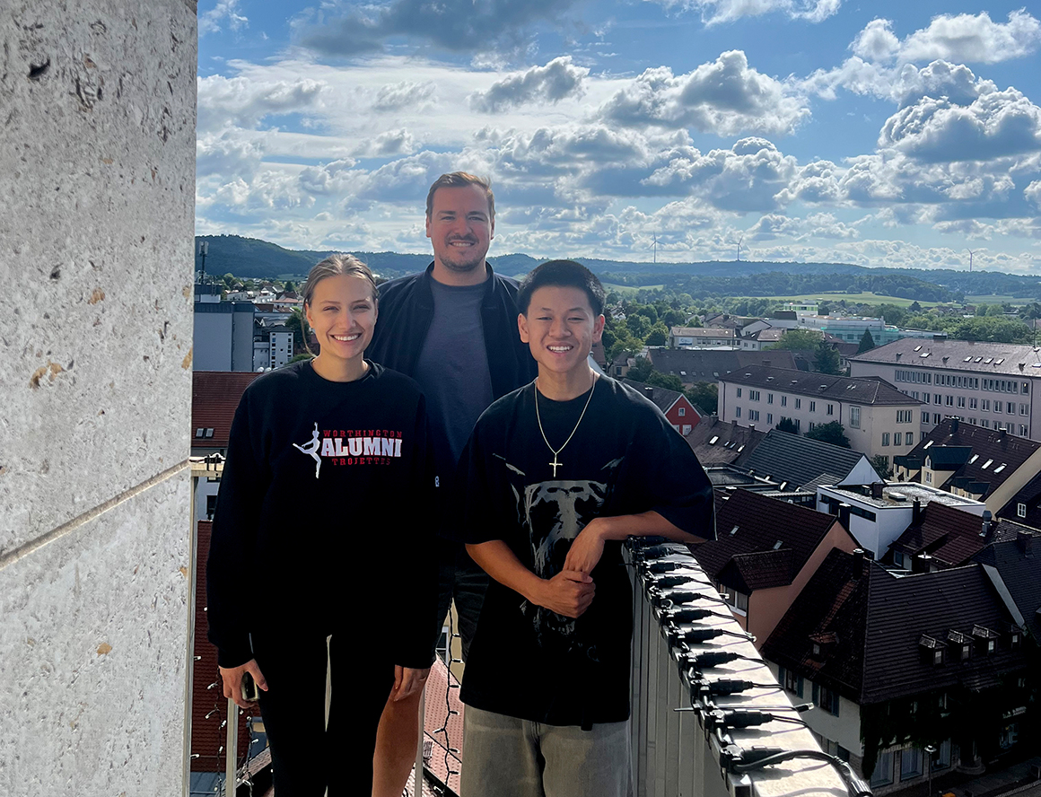drei Personen stehen lachend an einer Brüstung auf einem Turm, dahinter ein bewölkter Himmel