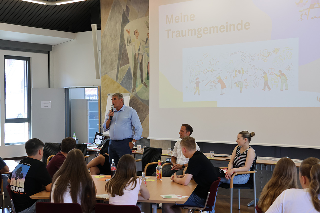 Ein älterer Mann mit Mikrofon in der Hand steht vor einigen Leuten