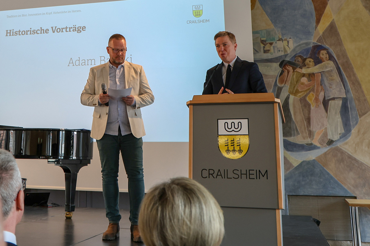 Zwei Männer auf einer Bühne, de reine hinter einem Rednerpult, der andere hat Zettel in der Hand.