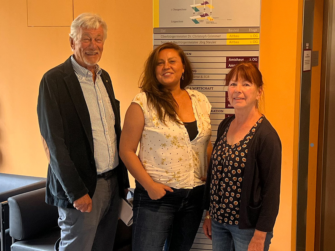 Ein Mann und zwei Frauen stehen vor einem Hinweisschild einer Verwaltung und lächeln in die Kamera